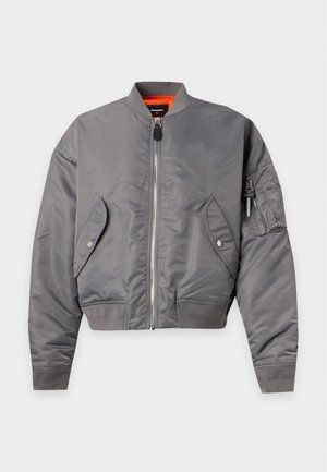 Homme portant une veste bomber grise, une chemise rouge, un jean bleu large avec des patchs et des baskets blanches, debout devant un fond clair uni.