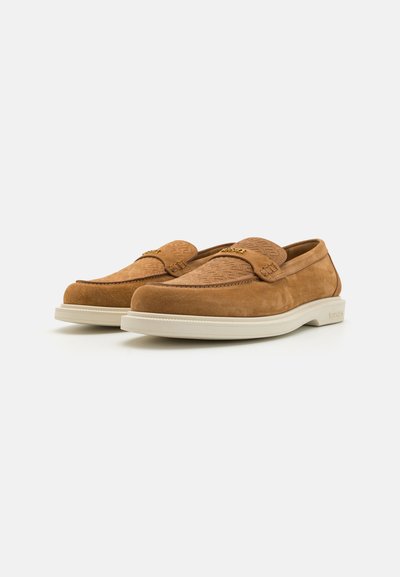 Tan suede loafers with a textured upper design and a white rubber sole. Features a small emblem on the strap and a smooth finish.