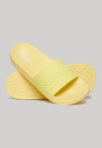 Yellow slide sandals with a textured rubber sole and a wide strap featuring embossed "SUPERDRY" branding. Smooth, matte finish.