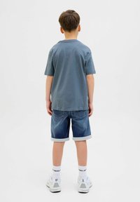 Un garçon portant un T-shirt bleu à manches courtes, un short en denim avec des poignets retroussés, des baskets blanches et des chaussettes avec des lettres noires, vu de dos.
