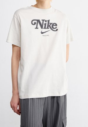 Personne portant un t-shirt blanc Nike avec le texte "Depuis 1972" et le logo swoosh noir, associé à un pantalon gris à fines rayures.
