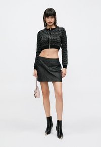 Pull noir court avec un motif, associé à une mini-jupette en denim foncé. Le mannequin porte des bottines noires et tient un petit sac à main rose.