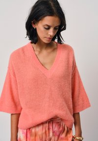 Zachte, oversized v-hals trui in licht koraal, gemaakt van donzig materiaal met korte mouwen; gedragen met gemusterde roze tie-dye broek.