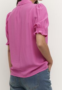 Blusa rosa de manga corta con cuello. Presenta mangas abullonadas con fruncido y una textura de tela suave. Combinada con jeans azul.