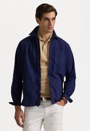 Dunkelblau, leichte Jacke mit durchgehendem Reißverschluss, spitzem Kragen und Brusttasche, darüber eine beige Poloshirt und eine weiße Hose.