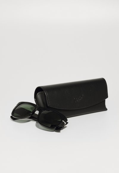 Črne sončne očala z zelenimi lečami, s sijočim plastičnim okvirjem in srebrnimi detajli, ob črni usnjeni torbici s pokrovčkom.