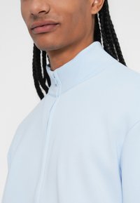 Sweatshirt azul-claro com fecho éclair, gola alta e textura suave, apresentado sobre um fundo branco.
