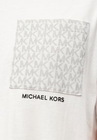 Λευκό μπλουζάκι με τσέπη από γκρι ύφασμα που διαθέτει επαναλαμβανόμενο μοτίβο "MK". Το "MICHAEL KORS" είναι κεντημένο με μαύρα γράμματα κάτω από την τσέπη.