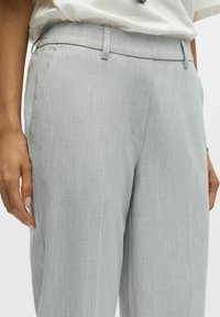 Zbliżenie na osobę w jasnoszarych spersonalizowanych spodniach z bocznymi kieszeniami i włożonej białej koszuli, z rękami swobodnie opadającymi wzdłuż ciała.