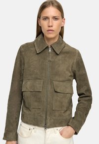 Oliwkowa zamszowa kurtka z zamkiem błyskawicznym, dwoma przednimi kieszeniami i kołnierzem do opadania. Gładka tekstura, skrócony krój.