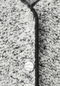 Tkanina crno-bijele boje s uzorkom pepita, naglašena srebrnim, teksturiranim gumbom i kontrastnim crnim obrubom.