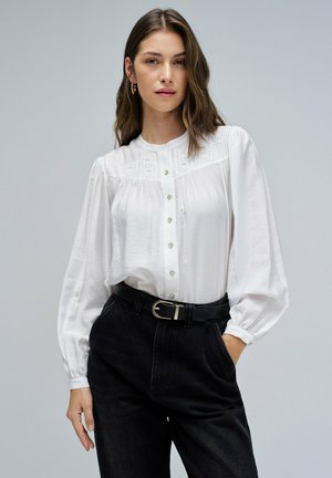 Blusa blanca con detalles de encaje y cierre de botones, combinada con pantalones oscuros de denim de tiro alto y un cinturón negro con hebilla plateada.