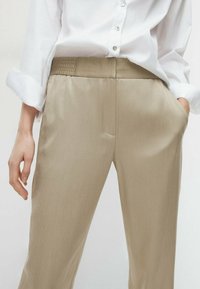Kobieta w beżowych, wysokich spodniach z elastycznym paskiem oraz białej koszuli zapinanej na guziki, z jedną ręką w kieszeni, widoczna od pasa do uda.