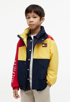 Junge trägt eine farbblockierte Jacke mit roten, gelben und marineblauen Paneelen, mit Tommy Hilfiger-Logo auf Brust und Ärmel.