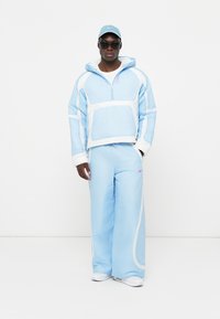 Jasnoniebieski dres z białymi akcentami. Bluza z zamkiem błyskawicznym i luźne spodnie, oba z miękką teksturą. Uzupełniony pasującą czapką.