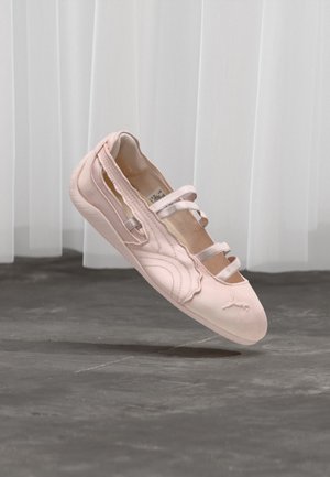 Light pink ballet flat with three elastic straps, scalloped edge, and rubber sole, suspended above a concrete floor with white curtains in background.