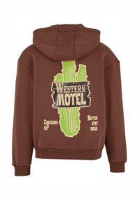 Ruda kapsulė su žaliu kaktuso grafiku ir tekstu "VAKARŲ MOTELIS". Įtraukta tekstas "Registruojatės?" ir "Geriau išlikite laukiniai." Minkšta medžiaga.