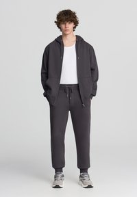 Felpa zip grigia scura con tasca a kangaroo, abbinata a pantaloni della tuta coordinati con vita e polsini elastici, indossati con scarpe da ginnastica.