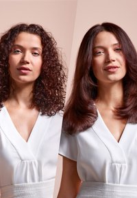 Twee vrouwen naast elkaar. Links: krullend, schouderlang, donkerrood haar. Rechts: steil, schouderlang, donkerbruin haar. Beiden dragen witte tops met V-hals.
