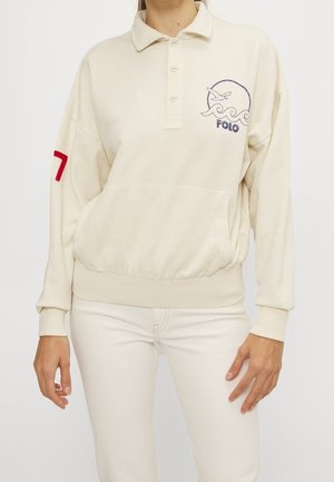 Kvinde iført en cremefarvet langærmet polotrøje med en måge- og bølgelogo, rød nummer 7 på ærmet, kombineret med cremefarvede bukser.