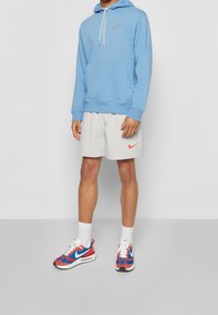 Ljusblå huvtröja med dragsko och framficka, i kombination med ljusgrå shorts med ett Nike-logotyp. Röda och blå Nike-sneakers fulländar looken.