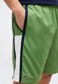 Πράσινο αθλητικό σορτς με μπλε σκούρες και λευκές ρίγες στο πλάι, ελαστική μέση, τσέπη με χέρι μέσα και το λογότυπο κροκόδειλου της Lacoste κοντά στο τελείωμα.