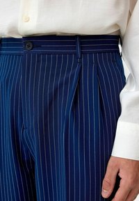 Marineblauwe gestreepte broek met een geplooide voorkant, knoopsluiting en riemlussen. Gemaakt van een gladde stof met fijne witte strepen.