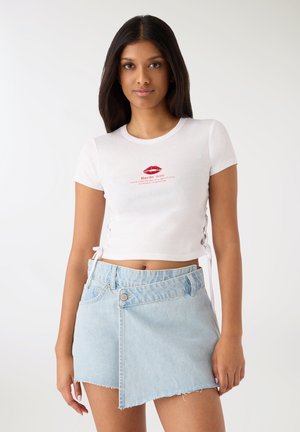T-shirt bianca in cotone con maniche corte e grafica di labbra rosse. Abbinata a una gonna in denim azzurro chiaro con orlo sfrangiato e design asimmetrico.