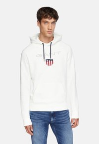 Weißer Hoodie aus weichem Stoff, mit einer Fronttasche, Kordeln und einem Logo im Schilddesign in Rot, Weiß und Blau.