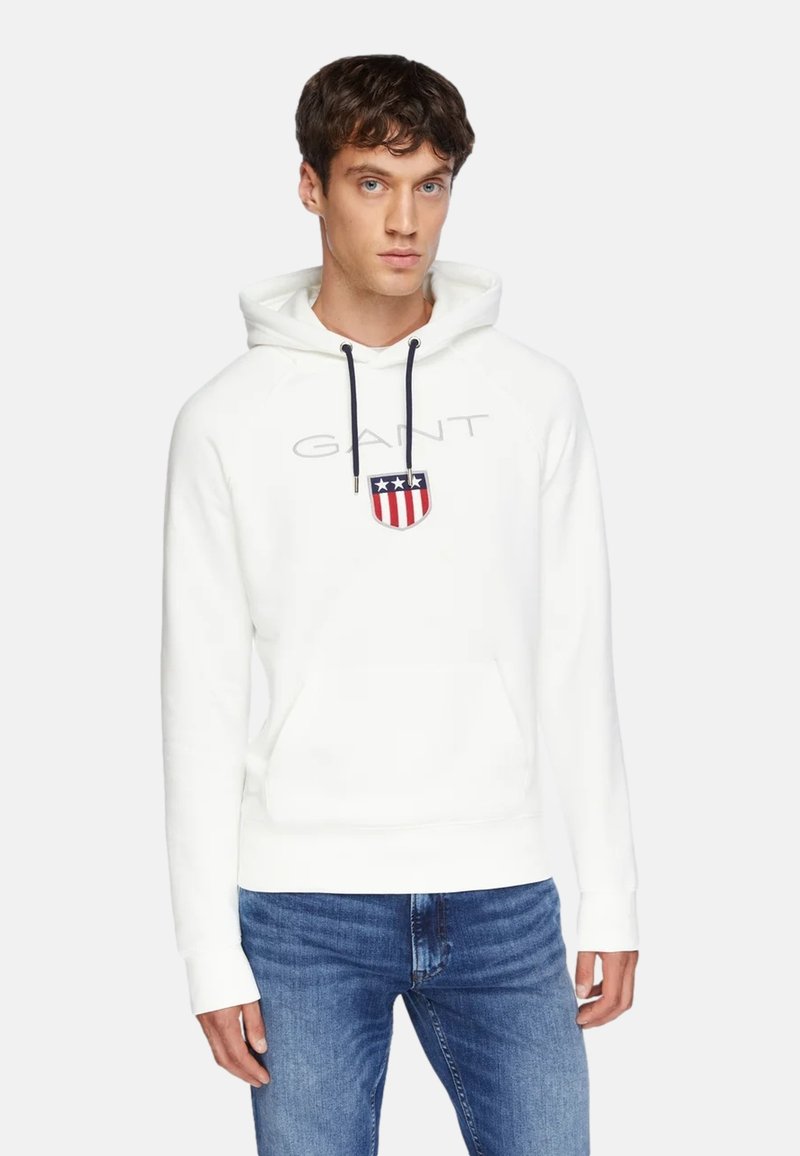 Weißer Hoodie aus weichem Stoff, mit einer Fronttasche, Kordeln und einem Logo im Schilddesign in Rot, Weiß und Blau.