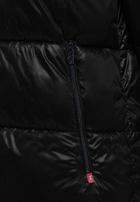 Czarna kurtka puchowa z błyszczącego materiału, posiada boczny zamek w kolorze granatowym, teksturowane panele oraz małą czerwoną metkę logo blisko zamka.