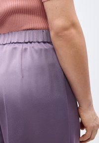 Κοντινό πλάνο ενός ατόμου που φοράει ριμπ ροζ τοπ και σατέν παντελόνι λεβάντας με ψηλή μέση, δείχνοντας το δεξί του χέρι και το πλάι.