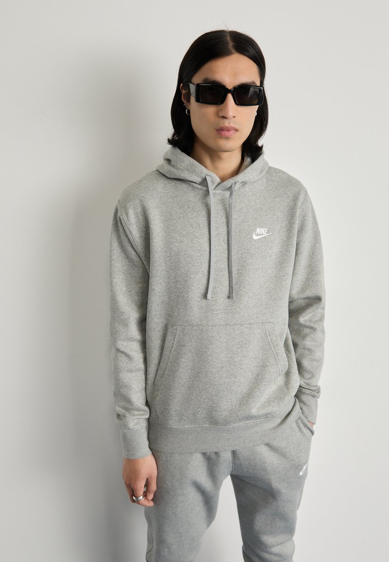 Grauer Nike-Hoodie mit Fronttasche, Kapuze mit Zugband und kleinem Logo. Kombiniert mit passenden grauen Jogginghosen. Glatte Textur, lässiges Design.