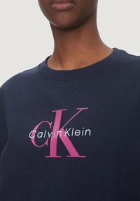 Marinblå crewneck sweatshirt med en stor fram-logos i rosa och vitt. Tyget verkar slätt med en standard passform och ribbad halsringning.