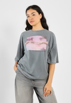 Gray oversized T-shirt with a loose fit, featuring a central print of hands against a pink and blue background; soft fabric texture.