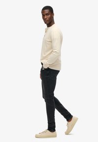 Chemise beige à manches longues avec trois boutons, associée à un jean foncé et des baskets beiges. Le modèle se tient de profil, les mains dans les poches.