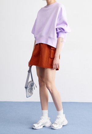 Henkilö pukeutuneena laventelinväriseen ylipitkään collegepuseroon, ruosteenväriseen taskulliseen hameeseen, valkoisiin sukkiin ja paksuihin valkoisiin tennareihin, pitäen kädessään pientä harmaata olkapussukkaa.