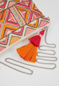 Pochette brodée ornée de motifs géométriques en rose, orange et crème avec des accents métalliques. Comprend une chaîne argentée amovible et un pompon décoratif rouge et orange.
