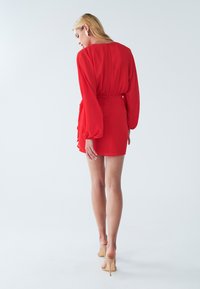 Rode jurk met lange mouwen, een getailleerde taille en een gerimpeld detail aan de zijkant. Gladde stof, tot boven de knie, gecombineerd met naaktkleurige hakken.