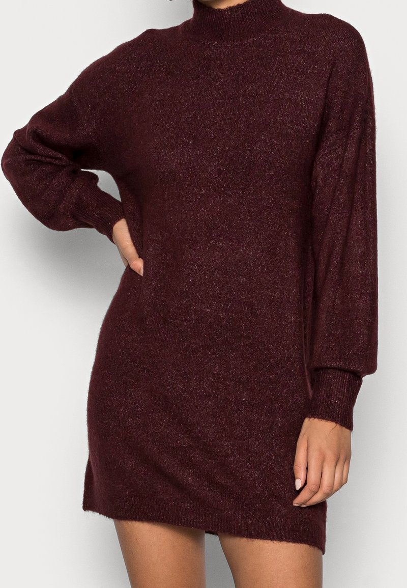 Robe-pull bordeaux en maille avec col montant, manches longues et coupe ample. Texture douce et poignets côtelés ajoutent du détail au design.