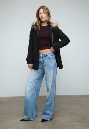 Kvinde iført en sort hættefrakke med pelskant, kort mørkelilla top, vidde lysblå jeans og sorte spidse sko står indendørs.