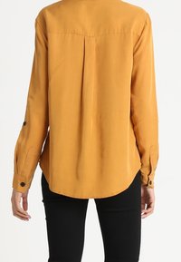 Senapsgul långärmad skjorta med rundad nederkant, detalj med plysch bak och knappdetaljer på ärmarna. Slät tygtextur.