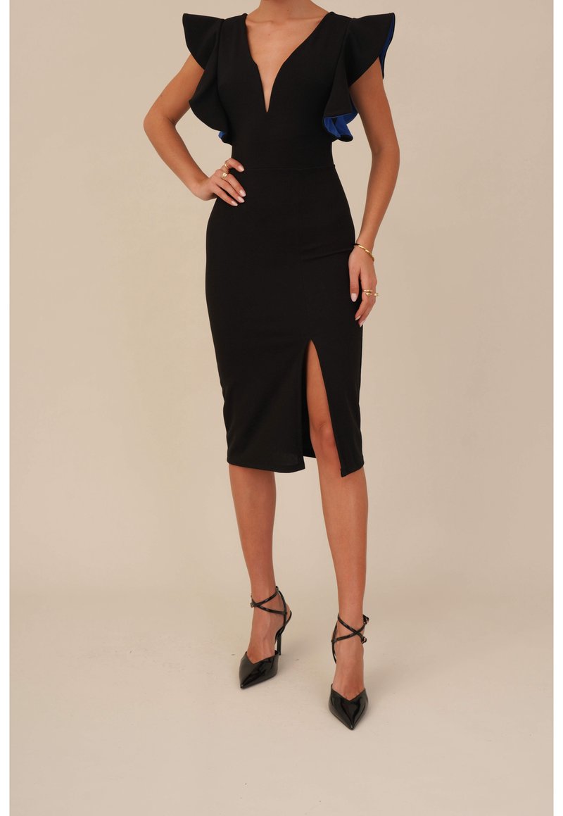 Femme portant une robe midi noire cintrée avec un décolleté en V profond, des manches à volants, une fente à la cuisse et des escarpins noirs pointus à talons hauts.