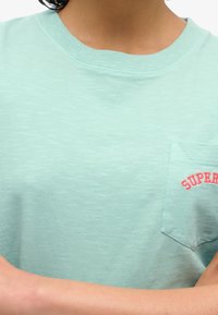 T-shirt in cotone verde menta chiaro con scollatura a girocollo, dotata di una tasca sul lato sinistro del petto e con la scritta "SUPER" ricamata in rosso su di essa.