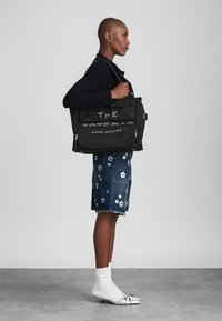 Czarna torba na ramię z płótna z białym nadrukiem "THE TOTE BAG MARC JACOBS." Posiada wiele kieszeni i pasek na ramię.