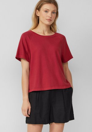 Rotes T-Shirt mit kurzen Ärmeln, Rundhalsausschnitt und lässiger Passform, kombiniert mit lockeren schwarzen Shorts. Die Materialien wirken weich und lässig.