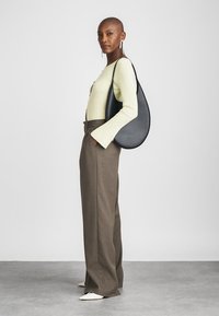 Melnas ādas plecu soma asaras formas, ar gludu tekstūru un minimālu aparatūru, valkāta ar gaismas zaļu ribotu topu un brūnām biksēm.