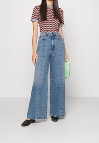 Donna che indossa jeans a vita alta e a gamba larga blu, una camicia a maniche corte con motivo ondulato rosa e nero, scarpe nere e tiene in mano una piccola borsa verde.
