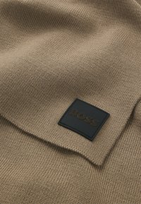 Strikket sweater i en lys brun nuance med en rektangulær sort læderlap, hvor "BOSS" er præget i brun.