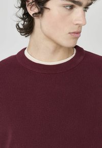 Pull en ribbed bordeaux avec un col rond, fabriqué en matériau tricoté texturé, superposé sur une chemise de base crème.